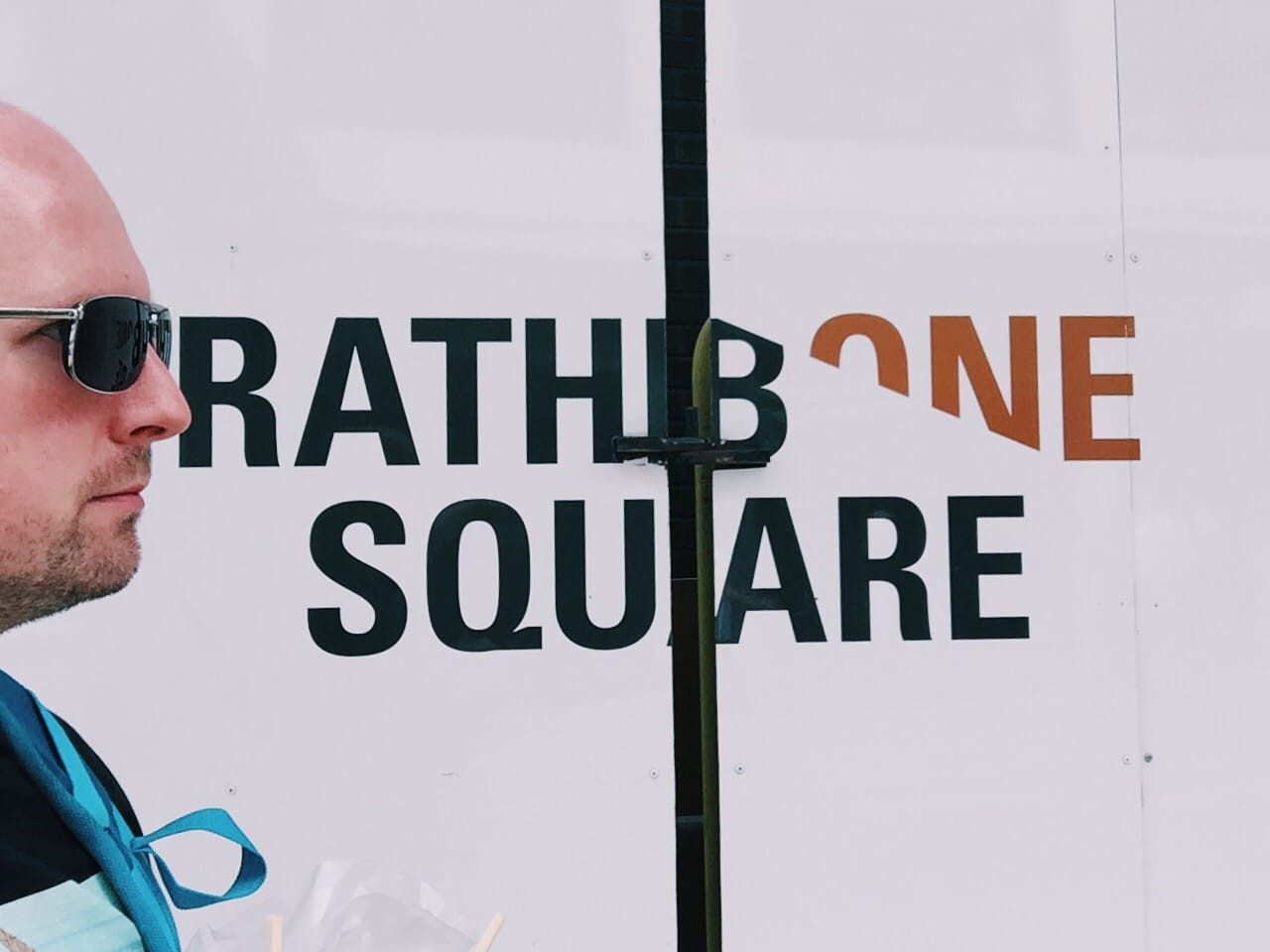 A bald man in sunglasses photographed in profile against a white wall bearing the words 'Rathbone Square' in large block lettering