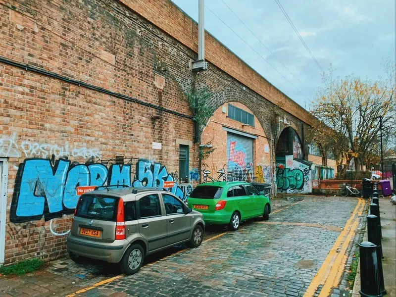 Two cars parked on a cobbled street beside a graffiti-covered Victorian railway arch in an urban area of London, on an overcast winter's day.