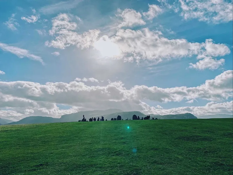 Visitors gathered around a stone circle on a vast green hillside under a bright, cloud-filled sky, with rolling fells in the distance.